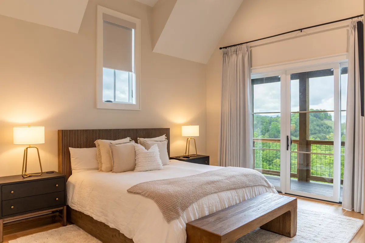 A cozy bedroom featuring a plush bed with decorative pillows, flanked by nightstands and softly lit lamps. The large windows flood the room with natural light and frame a scenic outdoor view, enhancing the suite's peaceful atmosphere.