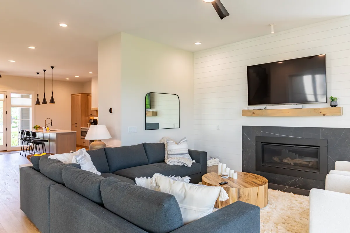 A cozy living room with a plush sectional sofa, a modern fireplace, a large TV, and a wooden coffee table. The space is designed for relaxation and socializing, enhanced by natural light streaming through large windows.