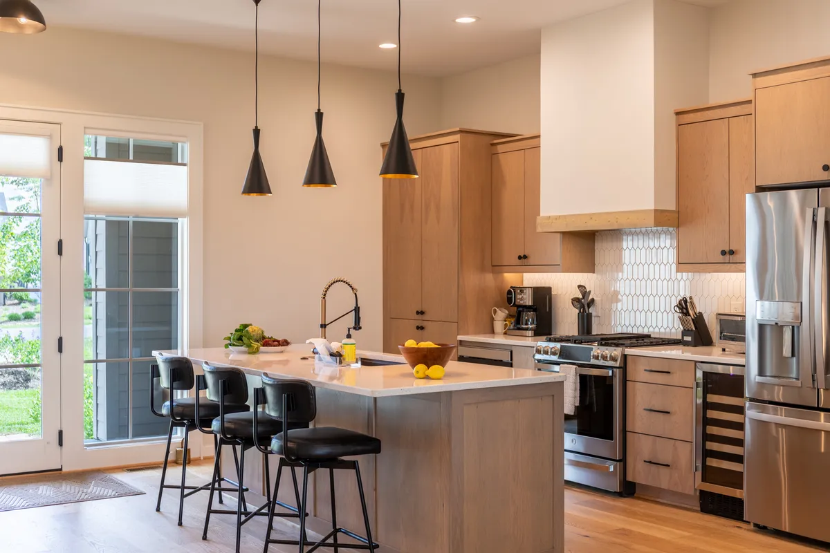 A bright, modern kitchen featuring a spacious quartz island with pendant lighting, stainless steel appliances, and a window offering views of the surrounding landscape. The island is surrounded by barstools, inviting social interaction and meal preparation. The kitchen seamlessly connects to adjoining living and dining spaces, conveying the open-concept layout.