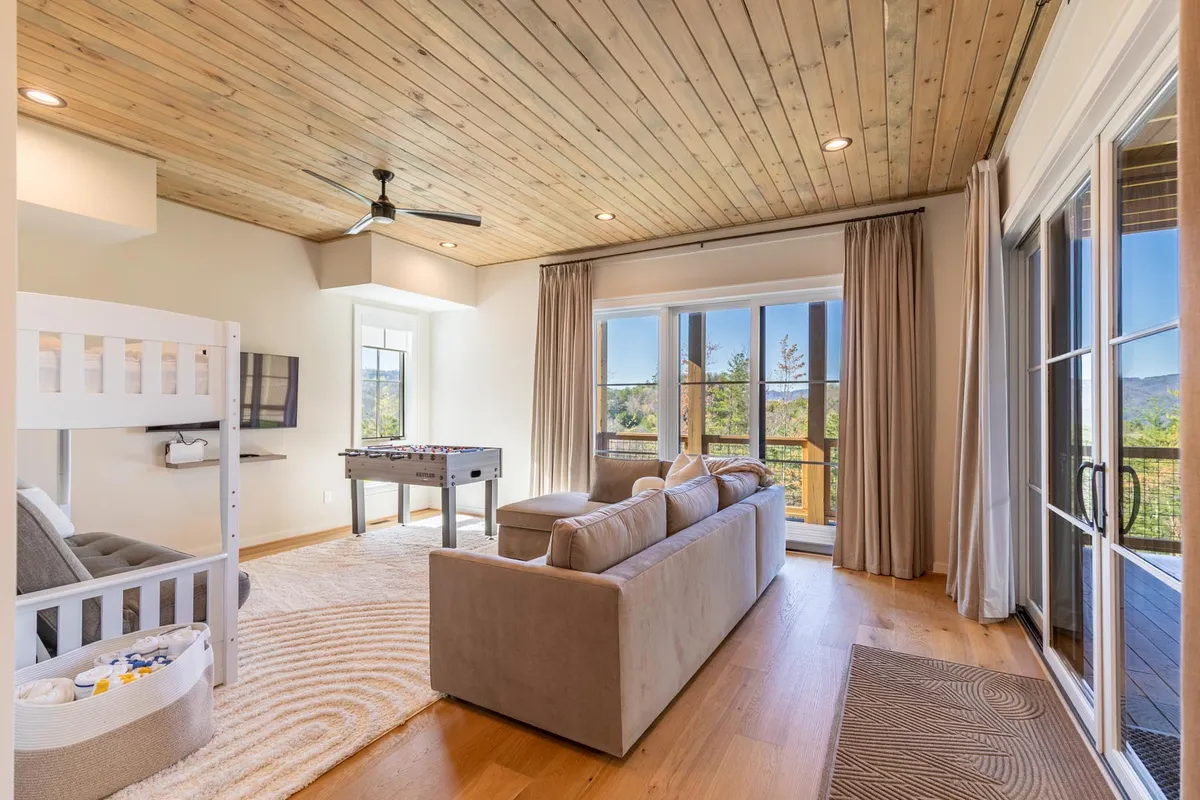 A bright, playful lower-level game lounge featuring a foosball table, cozy sofa, twin-over-futon bunk bed, and large windows showcasing soft natural light. The space is designed for fun and togetherness, with areas for gaming, relaxing, and reading.