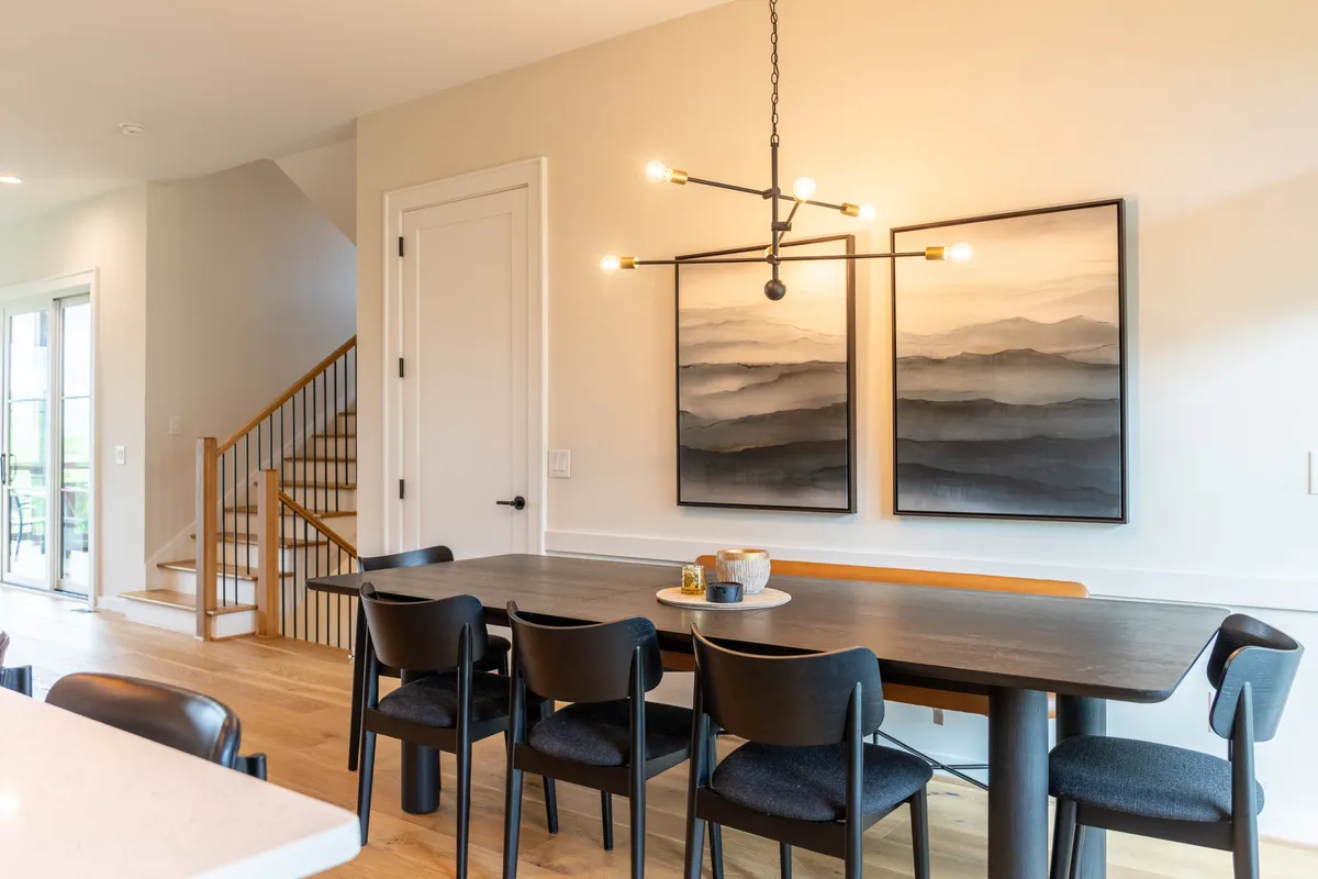 A contemporary dining area with a large table set for eight, positioned beneath a glowing chandelier. The space is adjacent to a staircase and features tasteful wall art, large windows, and stylish decor, making it ideal for family meals and social gatherings.