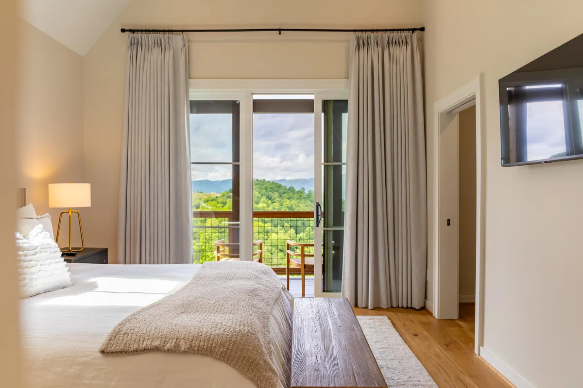 A serene bedroom with a large, comfortable bed facing a private balcony. The space is softly lit with natural light streaming through the curtains. The balcony reveals an expansive mountain view, inviting the outdoors in and creating a tranquil retreat.