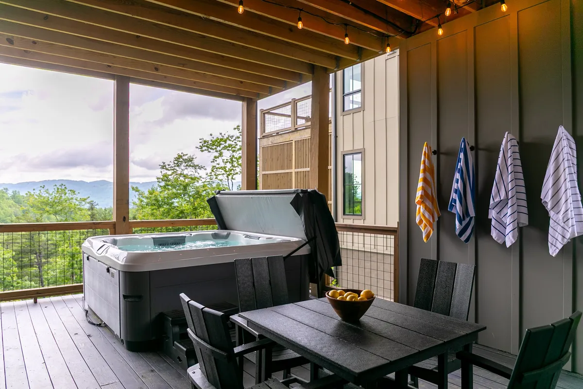 An inviting lower deck with a bubbling hot tub, dining table, and stunning mountain views in the background. Hanging towels and open space underscore the connection to nature and relaxation after a day of play.