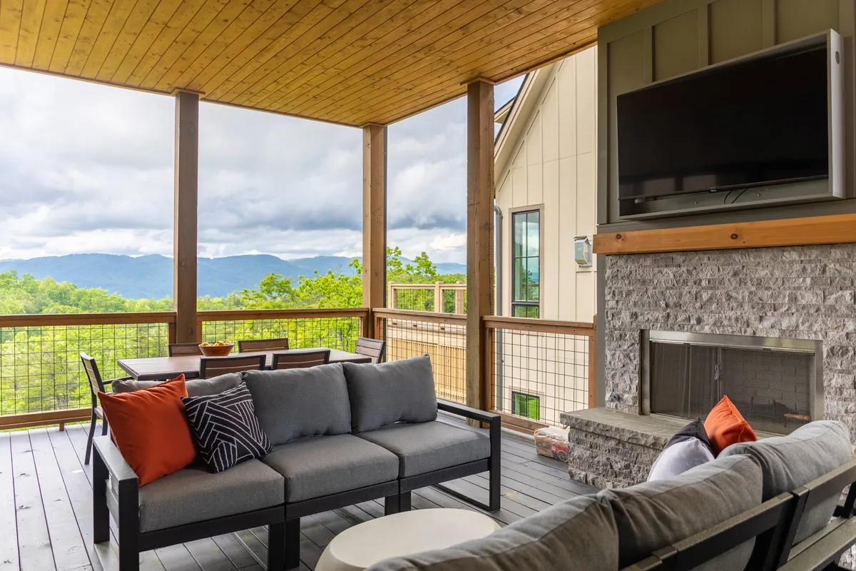 A main deck equipped with comfortable outdoor seating, a fireplace, and a television, all oriented toward expansive mountain views. The outdoor space blurs the boundary between inside and out, inviting al fresco dining and relaxation as the sun sets over the Blue Ridge Mountains.