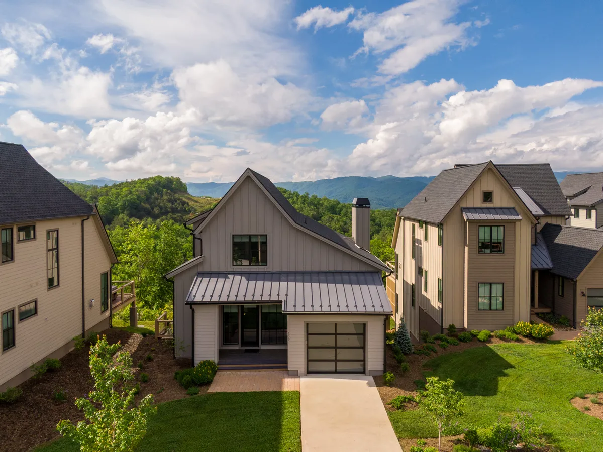 The image showcases the striking exterior of a modern single-family home set against the breathtaking backdrop of the Blue Ridge Mountains. The house features a welcoming porch, a beautifully landscaped yard, and expansive views of rolling peaks. The scene is bathed in golden light, suggesting a perfect moment for sunset gatherings or peaceful mornings.